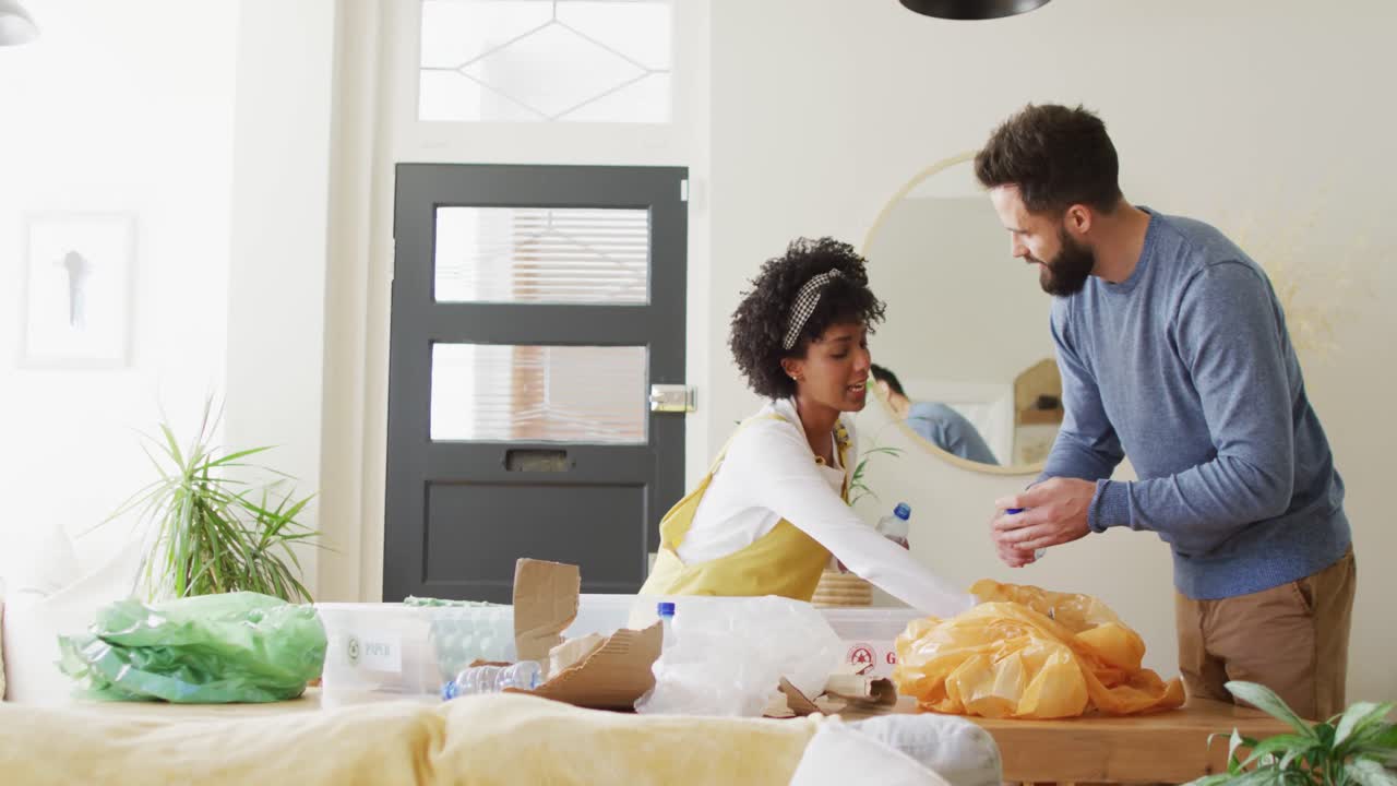 Video of happy diverse couple having fun sorting recycling at home, with copy space