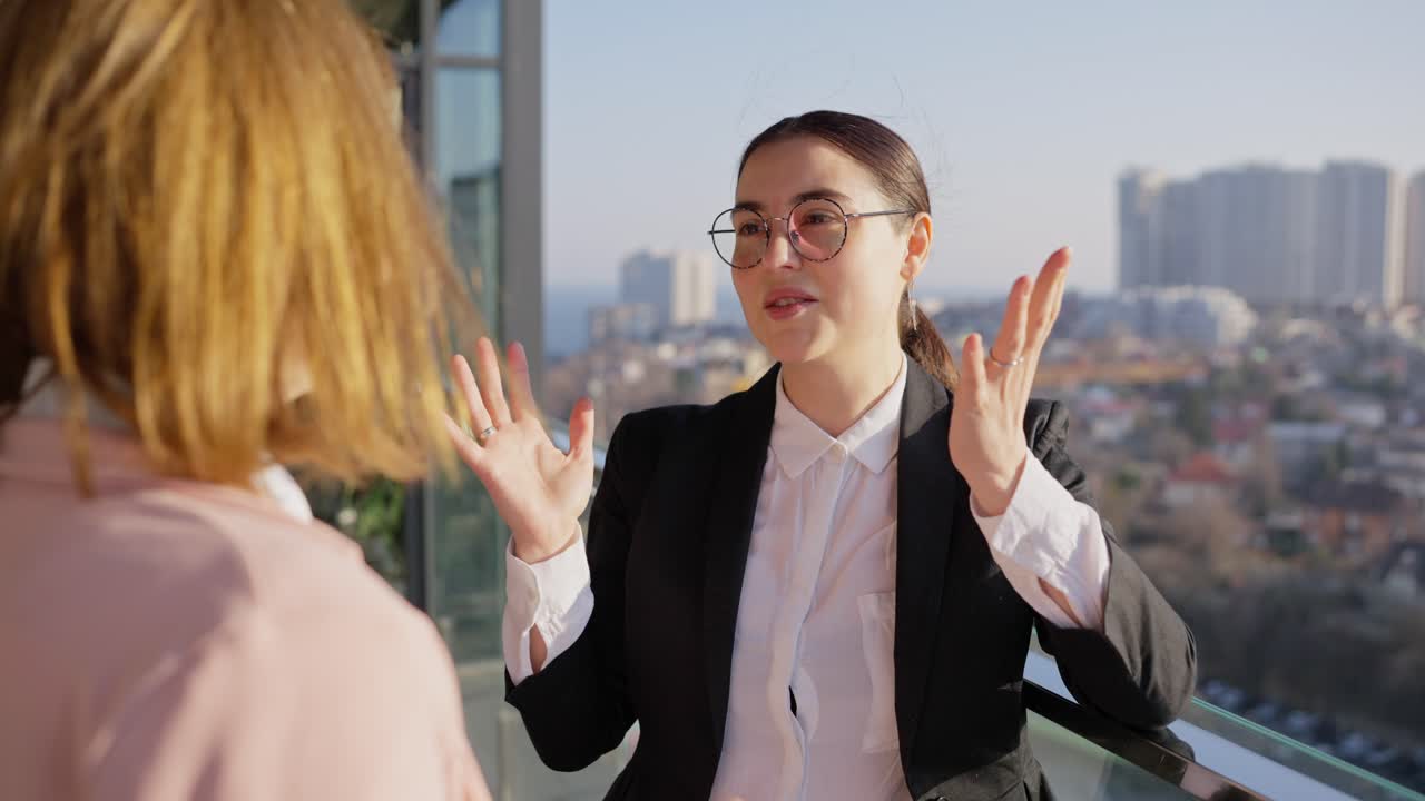 sobre el hombro una confiada chica morena con gafas redondas y un uniforme de negocios negro se comunica con su colega una chica rubia con un peinado bob mientras se relaja en la terraza mientras trabaja con vistas a la ciudad en un clima soleado por la noche