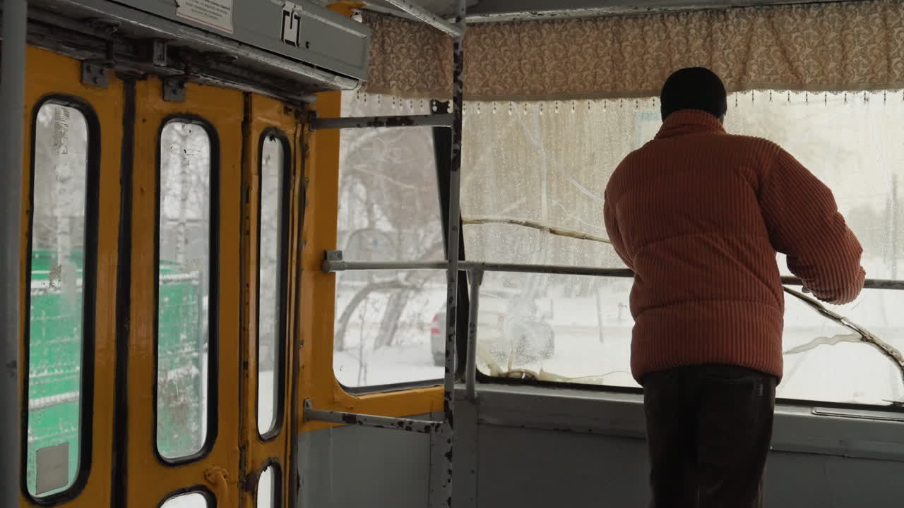 Uomo che ispeziona la porta del tram in mezzo alla neve, pannelli interni gialli incorniciano la scena, l'operatore si china verso il vetro smerigliato e la maniglia di metallo, auto parcheggiate e recinzione verde visibili all'esterno, atmosfera di manutenzione con attenzione