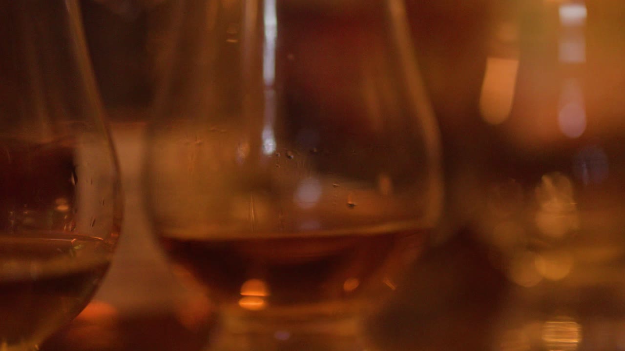 Una mano si allunga per prendere un bicchiere di whisky color ambra, illuminato da luci calde in un pub