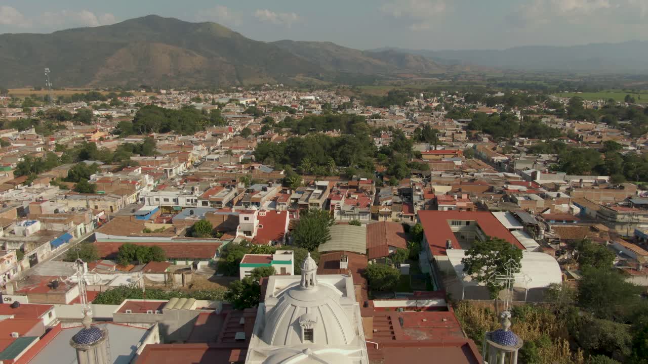 Aerial flyover church twin bell towers and rooftop, tilt up to reveal small Mexican town and mountain range in Jalisco, Mexico