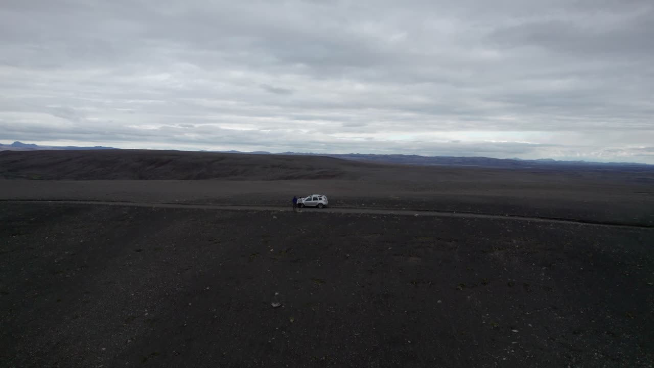 A couple of people stand on the side of a road in a remote Icelandic desert, surrounded by vast, barren landscapes. The empty road stretches through the rugged terrain under an open sky.