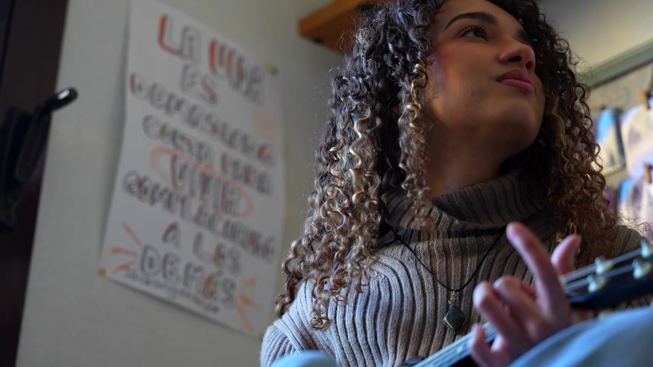 Donna latina con i capelli ricci che strimpella un ukulele mentre compone musica nel suo accogliente appartamento