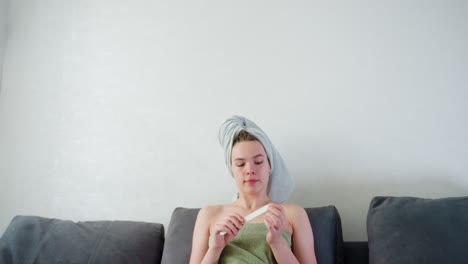 close view of young woman wrapped in light green towel with hair wrapped in white towel filing nails while seated on couch in bright indoor space, portraying peaceful post-bath selfcare moment