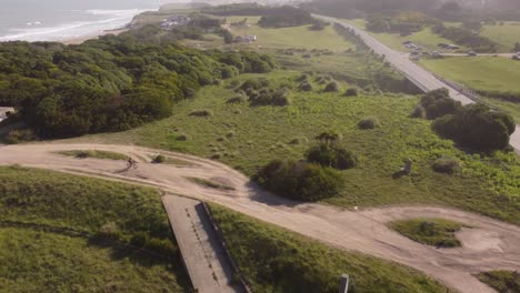 Cyclist-pedaling-along-dirt-road-with-ocean-in-background,-Chapadmalal