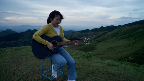 the tranquility of nature is complemented by the gentle strumming of a woman’s guitar as she sits on a hilltop, lost in her music with the mountains in the background