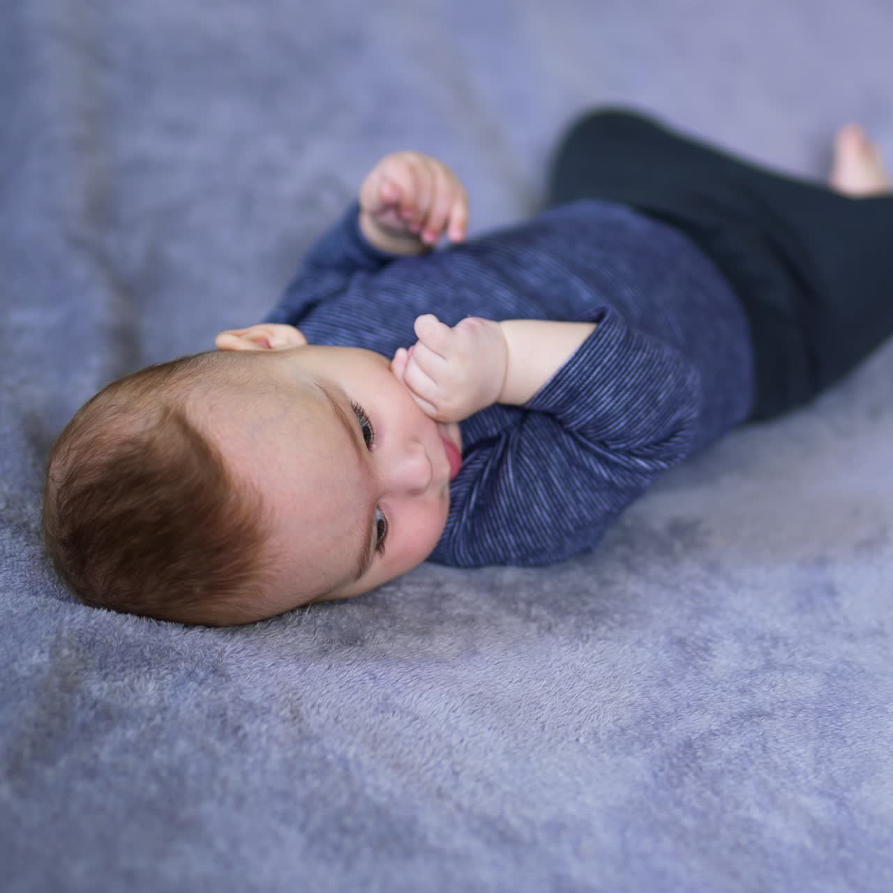 Calm little infant boy lying peacefully on his back. Beautiful child chewing his fingers and looking sideways. Blurred grey background