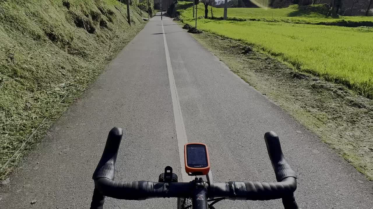 Visuale in prima persona (POV) da una bicicletta che percorre una strada rurale deserta attraverso una campagna verdeggiante.