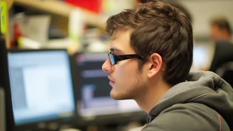 Young programmer wearing glasses looking at computer screen in an office environment, possibly reviewing code or working on a software development project