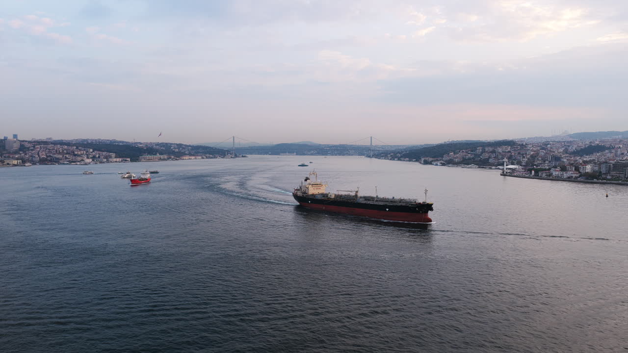 A cargo ship sails through the Bosphorus Strait at dusk, surrounded by Istanbul’s hilly coastline and distant bridges connecting Europe and Asia