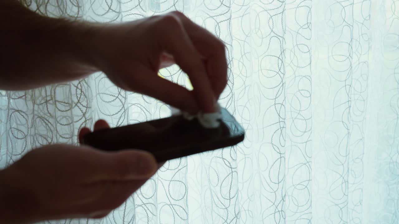 Bredt billede af hænder, der tørrer en smartphone-skærm af med et stykke papir foran et lyst vindue – ideelt til indhold om rengøring af teknologi, hygiejnerutiner og pleje af enheder