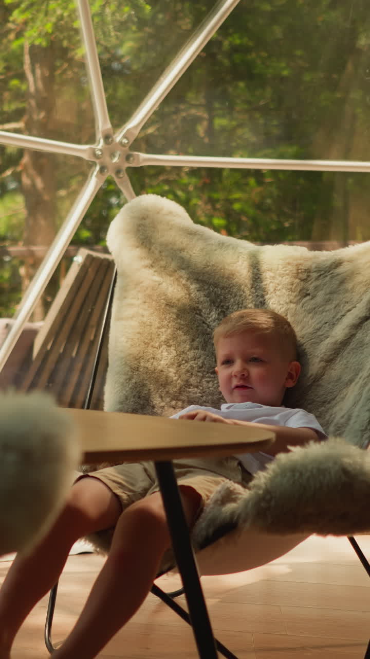 Younger brother talks to elder sister sitting in near furry chair with legs dangling. Children enjoy lazy day and beautiful forest outside window. Family vacation in mountains