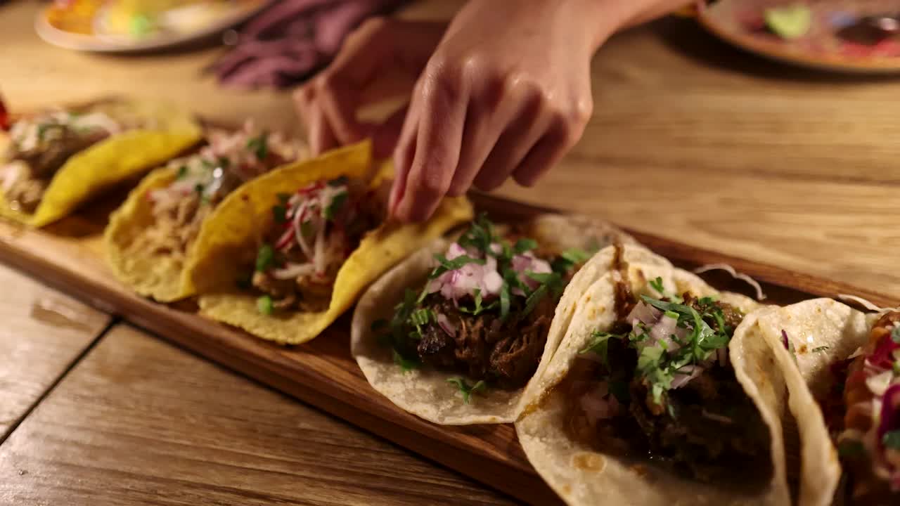 Multiple hands reach for assorted grilled tacos on a rustic wooden table under warm, ambient lighting, emphasizing communal dining and vibrant street food culture