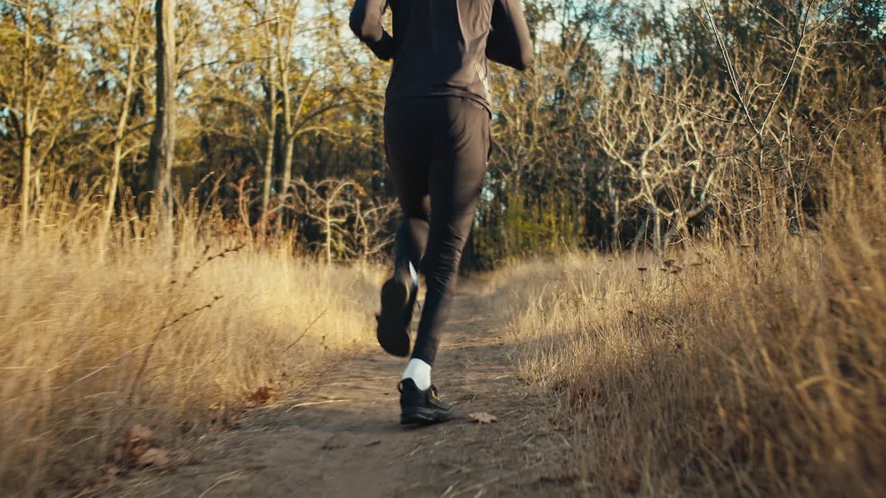vista trasera de un hombre en un uniforme deportivo negro en zapatillas negras corre a lo largo de un camino estrecho entre la hierba seca en el bosque de otoño por la mañana. correr por la mañana en el parque en otoño