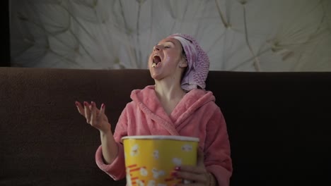 Woman-watching-a-late-night-movie-at-TV,-eating-popcorn.-Bathrobe,-facial-mask