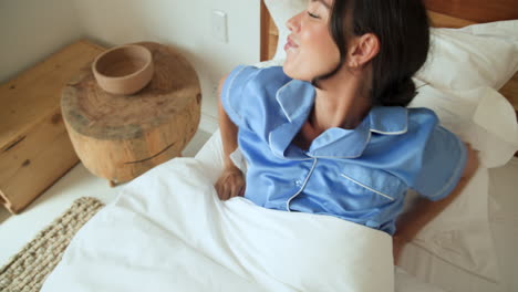 Woman in bed wearing blue pajamas, gently massaging temples, feeling relaxed, at home