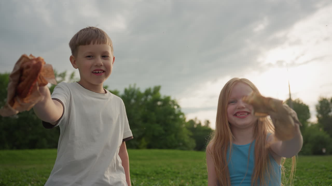 portrait of picnic children showing snacks playfully making funny faces in park green grass sunset background smiling holding pastries with hands celebrating summer outdoor family time
