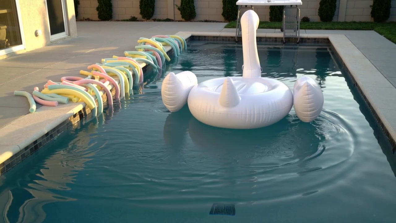 Drifting white swan float being pushed by current toward far home pool wall, passing pastel noodles