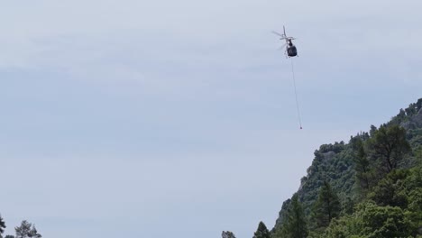 a helicopter emerges from behind forest terrain, carrying equipment to a remote, hard-to-reach location. aerial reveal of logistics in rugged natural landscape