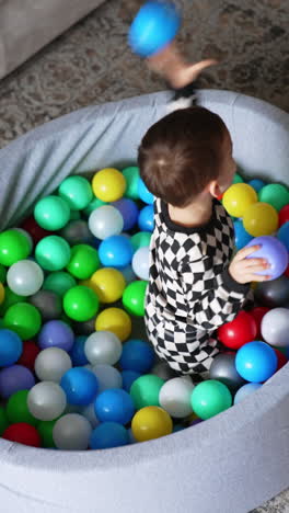 baby boy in black and white pajama sits in the dry pool. smiling playful kid throws the balls and tumbles in the pool.