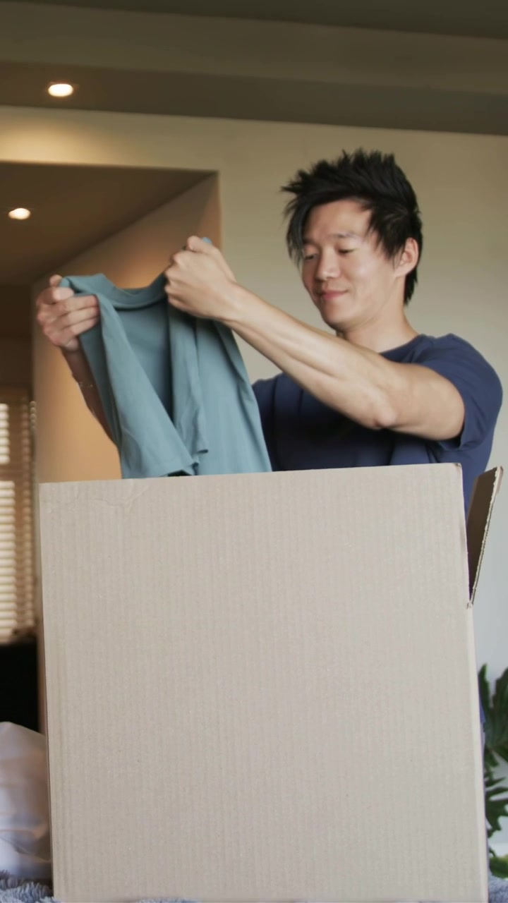 Vertical video: Reaching into box, man in navy tee unfolding, inspecting teal shirt on bed sorting