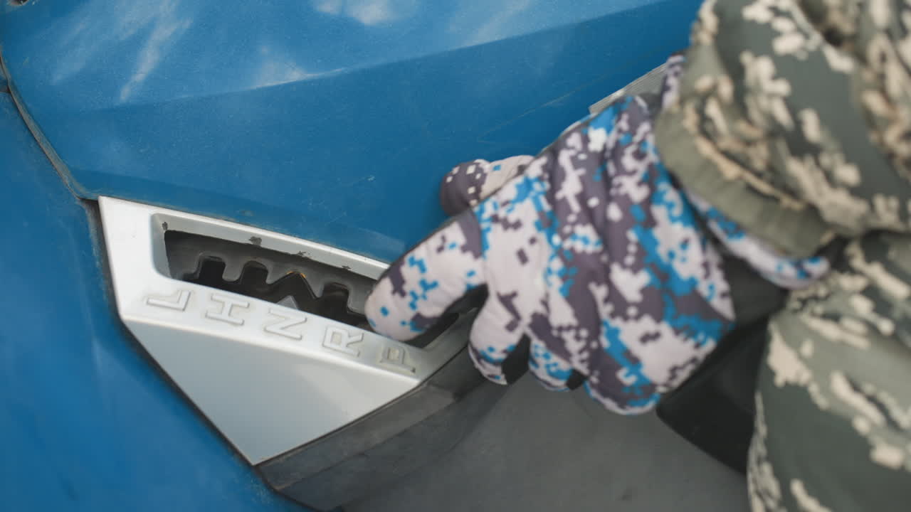 Main gantée ajustant le sélecteur de vitesse d'un quad bleu, manche de camouflage visible, séquence en gros plan montrant le mouvement du bouton et l'engagement du levier, contrôle tactile lors d'un contrôle d'entretien, environnement extérieur robuste.