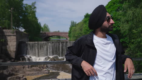 Punjabi-Sikh-Man-In-Sunglasses-And-Dastar-Turban-Standing-On-The-Bridge-With-Vanhankaupunkoski,-Old-Town-Waterfall-In-Helsinki,-Finland