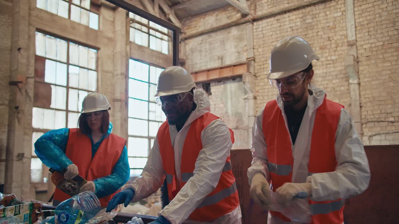 A trio of factory workers in special white uniforms and orange vests and white helmets recycle garbage and plastic bottles on a conveyor belt