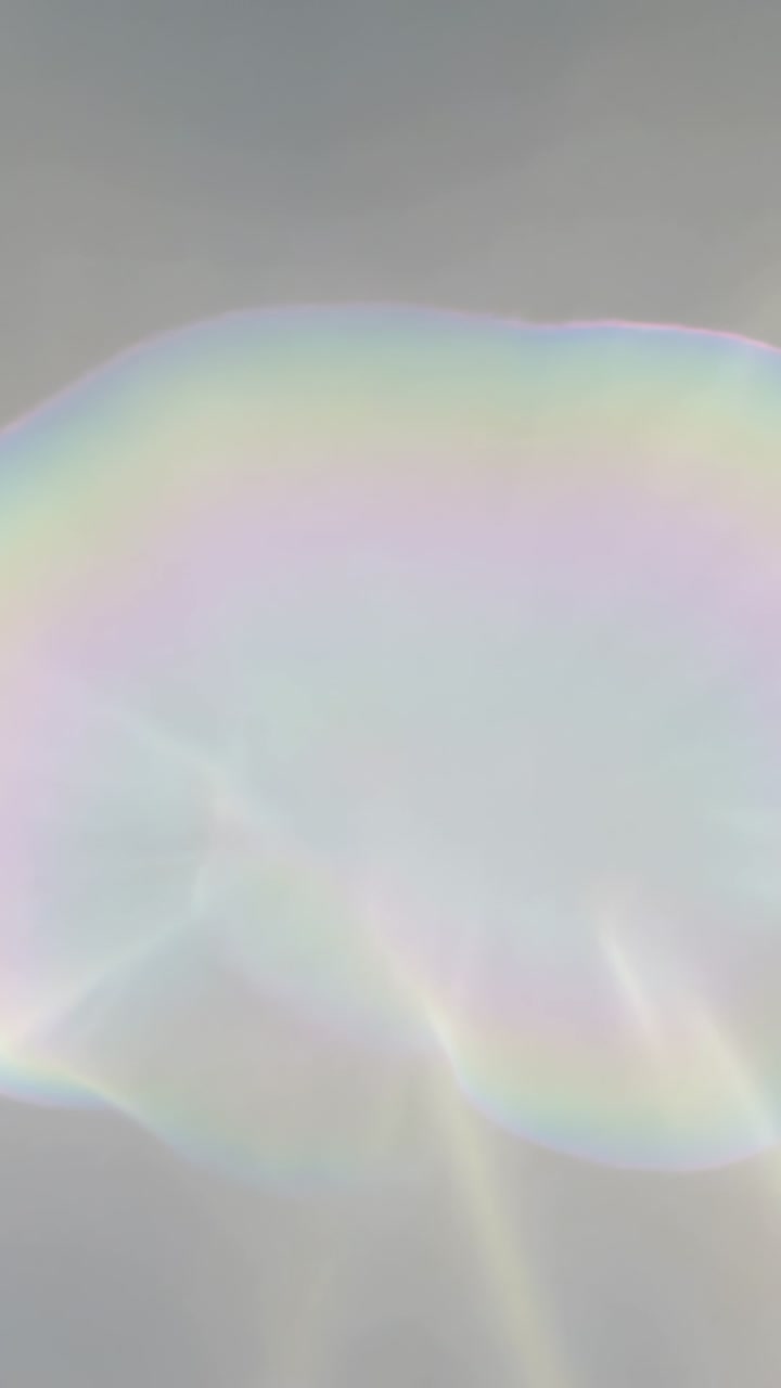 Vertical video: Shifting light moving iridescent film over pale gray backdrop, rainbow streaks
