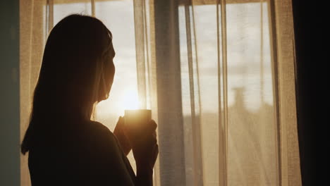 middle-aged woman with a cup of tea in her hands. looking out the window at sunset