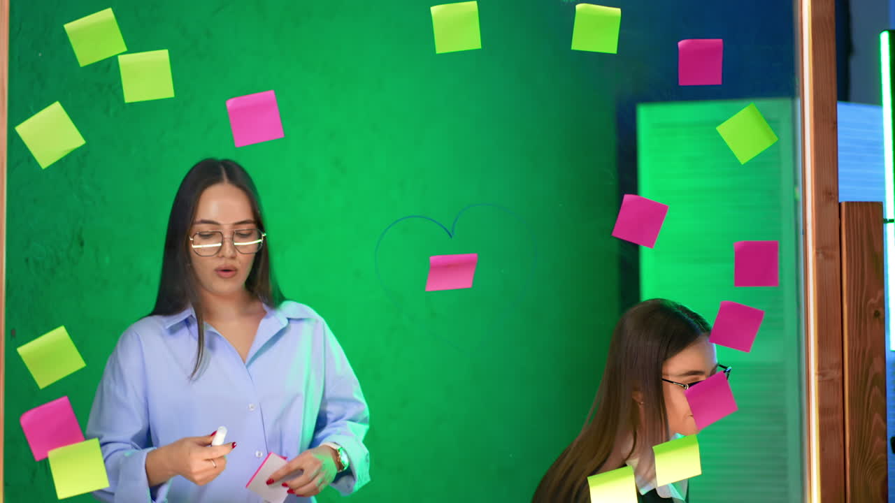 Cheerful ladies working in the office stand at the glass board. Woman in black jacket draws a heart around one sticky note.