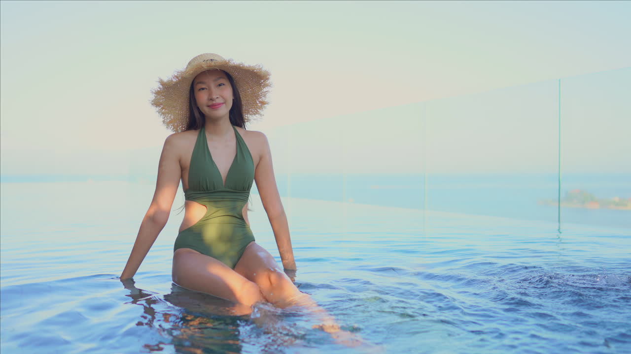 Pretty young Asian woman wearing green bathing suit and large straw hat sitting on edge of infinity pool kicking her feet in the warm water and smiling