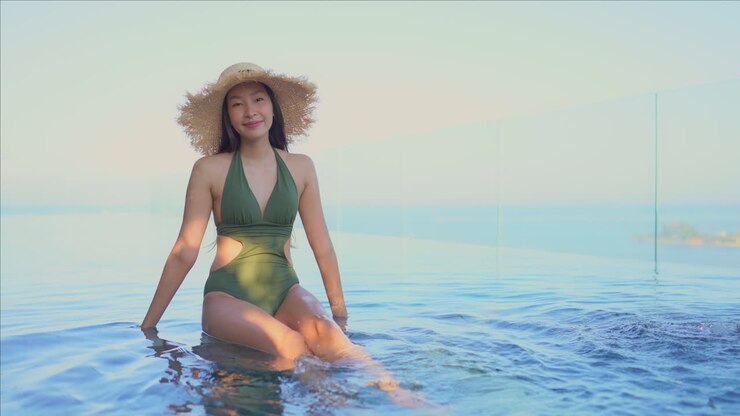 Pretty young Asian woman wearing green bathing suit and large straw hat sitting on edge of infinity pool kicking her feet in the warm water and smiling