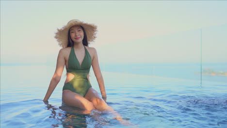 pretty young asian woman wearing green bathing suit and large straw hat sitting on edge of infinity pool kicking her feet in the warm water and smiling