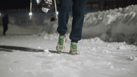 leg view of skater walking home on snowy ground at night, holding ice skates by laces with people blurred in background, wearing green sneakers and festive socks under dim winter lighting