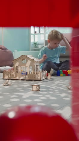 blond toddler plays with toys and blocks sitting on floor. little boy builds structures in living room view through clearance in construction of cubes