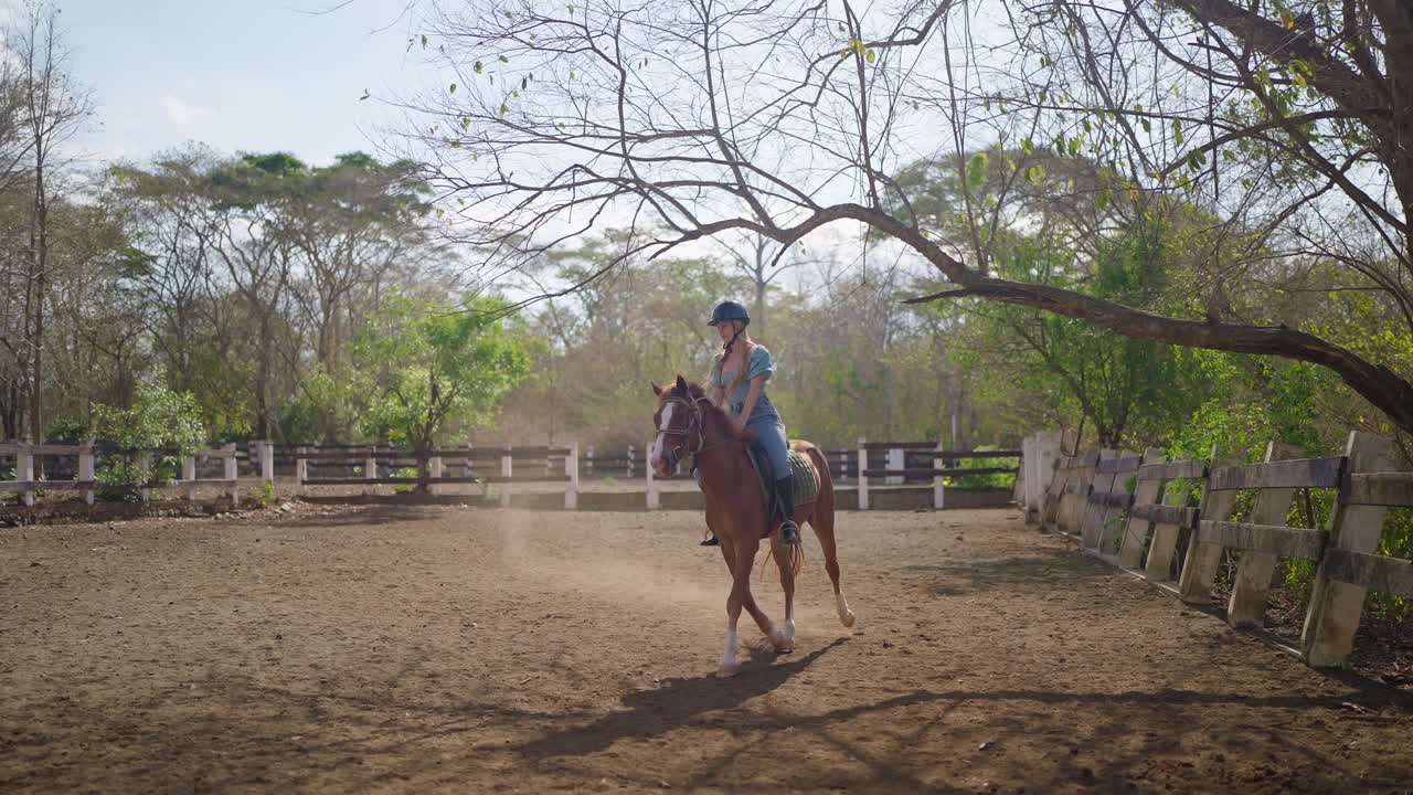Uma mulher cavalga um cavalo marrom em uma pista de corrida, usando capacete e jeans. Cercada por cercas brancas e chão de terra marrom, a cena captura o esporte equestre, movimento e a atmosfera de treinamento ao ar livre.