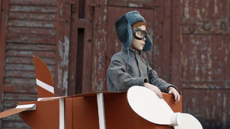 retrato de un lindo niño pelirrojo con sombrero y gafas parado en un avión de cartón y sonriendo a la cámara