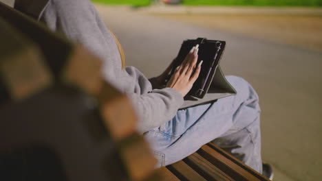 lady viewed from behind sitting on park bench at night, engaged with turned-off tablet, sliding through screen, legs crossed, phone in back pocket, illuminated by soft park lighting