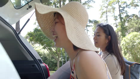 happy asian female friends putting suitcases into car trunk and laughing on sunny day, slow motion