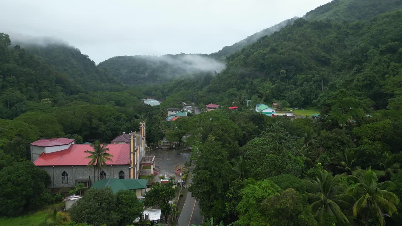 A wide aerial shot glides over a forested Philippine village. Colorful rooftops peek through dense greenery as mist rolls across the mountain slopes