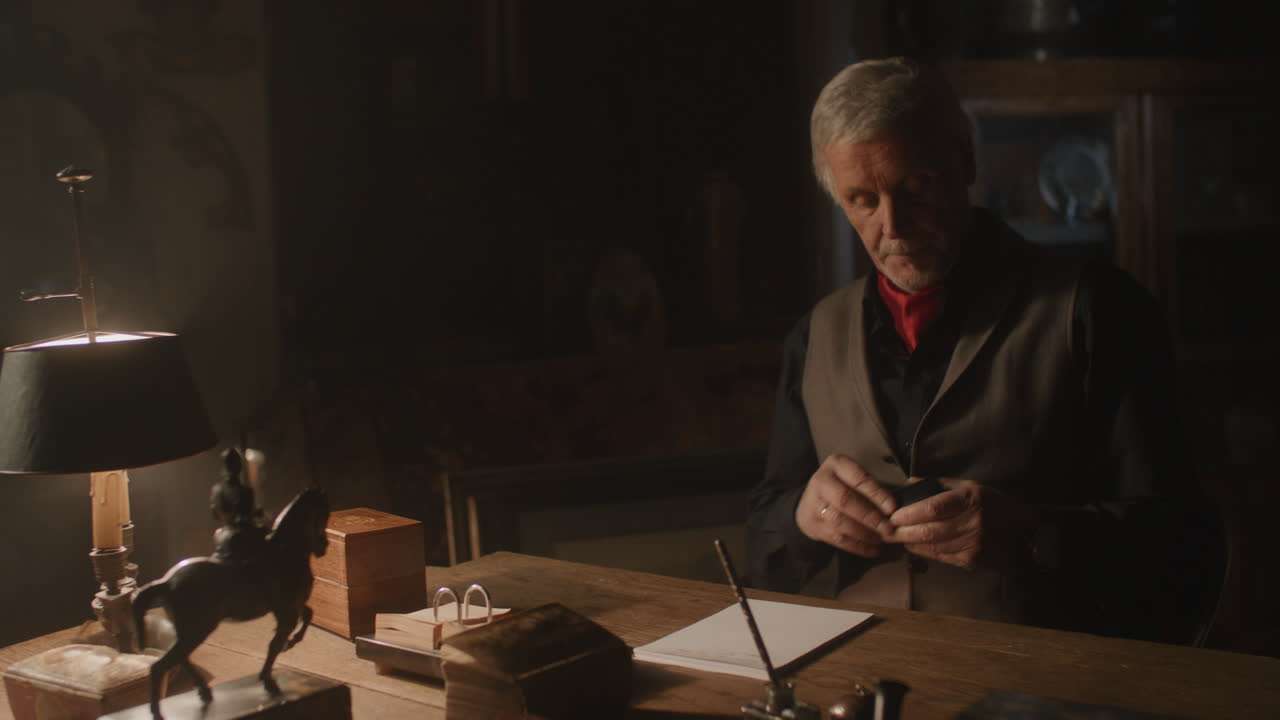 Old passionate man sits down to his desk, put on the glasses and writes a letter