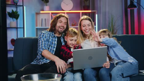 simpática sonrisa satisfecha familia joven sentada en el sofá suave en casa por la noche durante el chat de video con los parientes en la computadora portátil y saludándolos con las manos agitadas