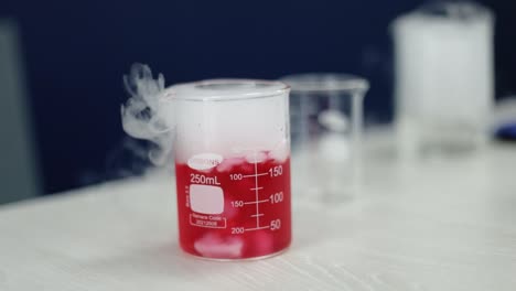 en sorprendente cámara lenta, un vaso de vidrio lleno de un líquido rojo luminoso sufre una transformación química, a medida que los zarcillos de humo blanco se elevan, encarnando la complejidad de las maravillas científicas