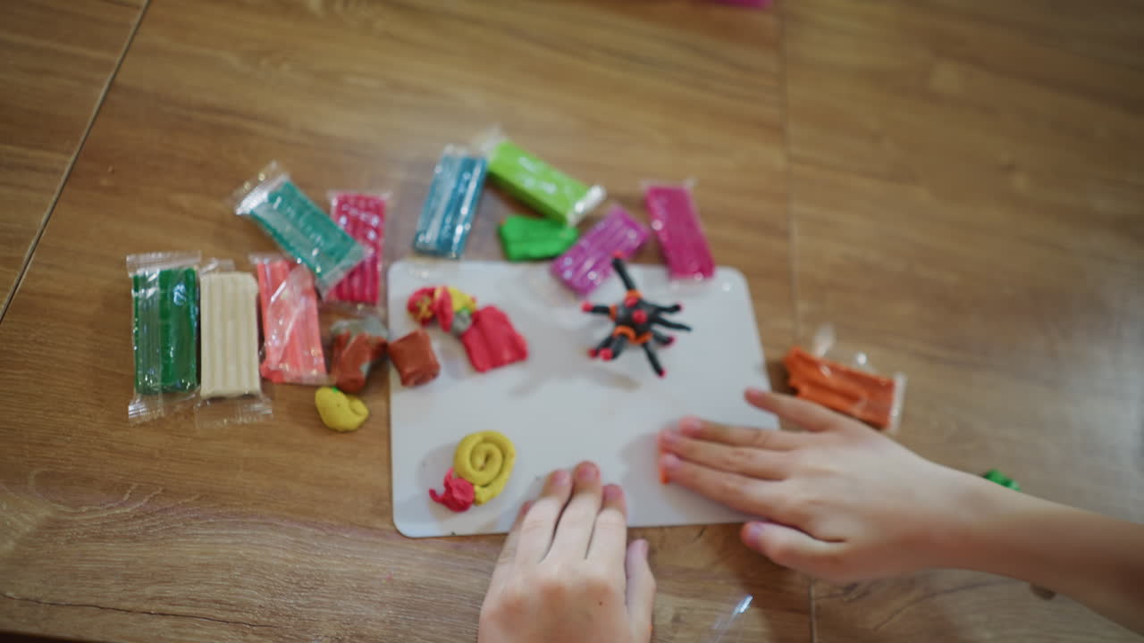 Hands of child shaping colorful modeling clay on white board, surrounded by packets of plasticine in multiple shades, creating imaginative figures with focus, creativity, expression at home