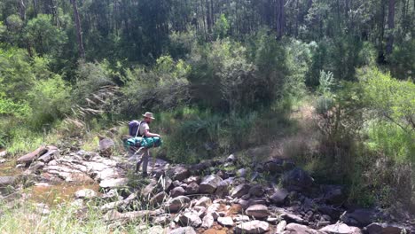 an australian bush man crosses a creek while going on walkabout with his swag