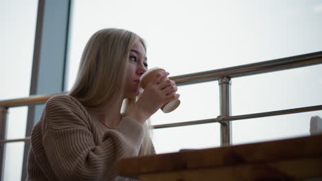 side view of lady sipping coffee while holding a disposable cup in cozy mall with iron frame window structure, modern mall ambiance with soft lighting, perfect for relaxation