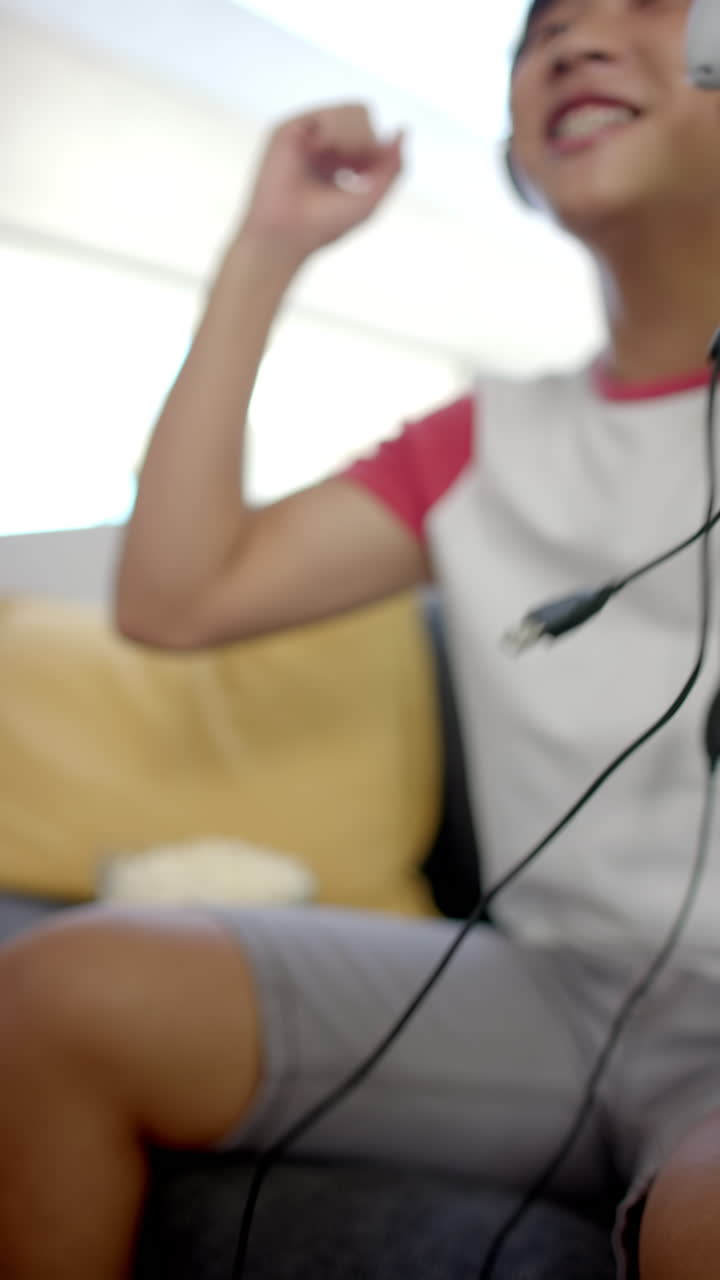 Vertical video: Celebrating victory, Asian teenage boy playing video game with headphones at home