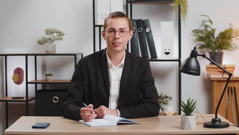 young businessman looking to camera and writing down on diary in video chat at home office desk