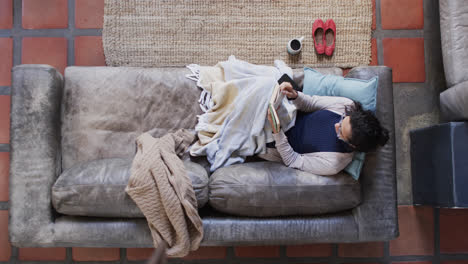 Vista-Aérea-De-Una-Mujer-Birracial-Tumbada-En-Un-Sofá-Bajo-Una-Manta-Y-Leyendo-Un-Libro-En-Casa,-Cámara-Lenta